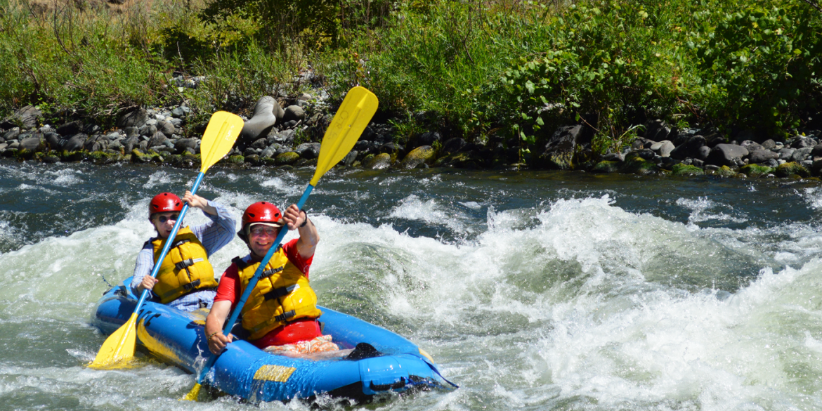 Two trip participants in a blue raft smiling as they navigate a river's whitewater rapids