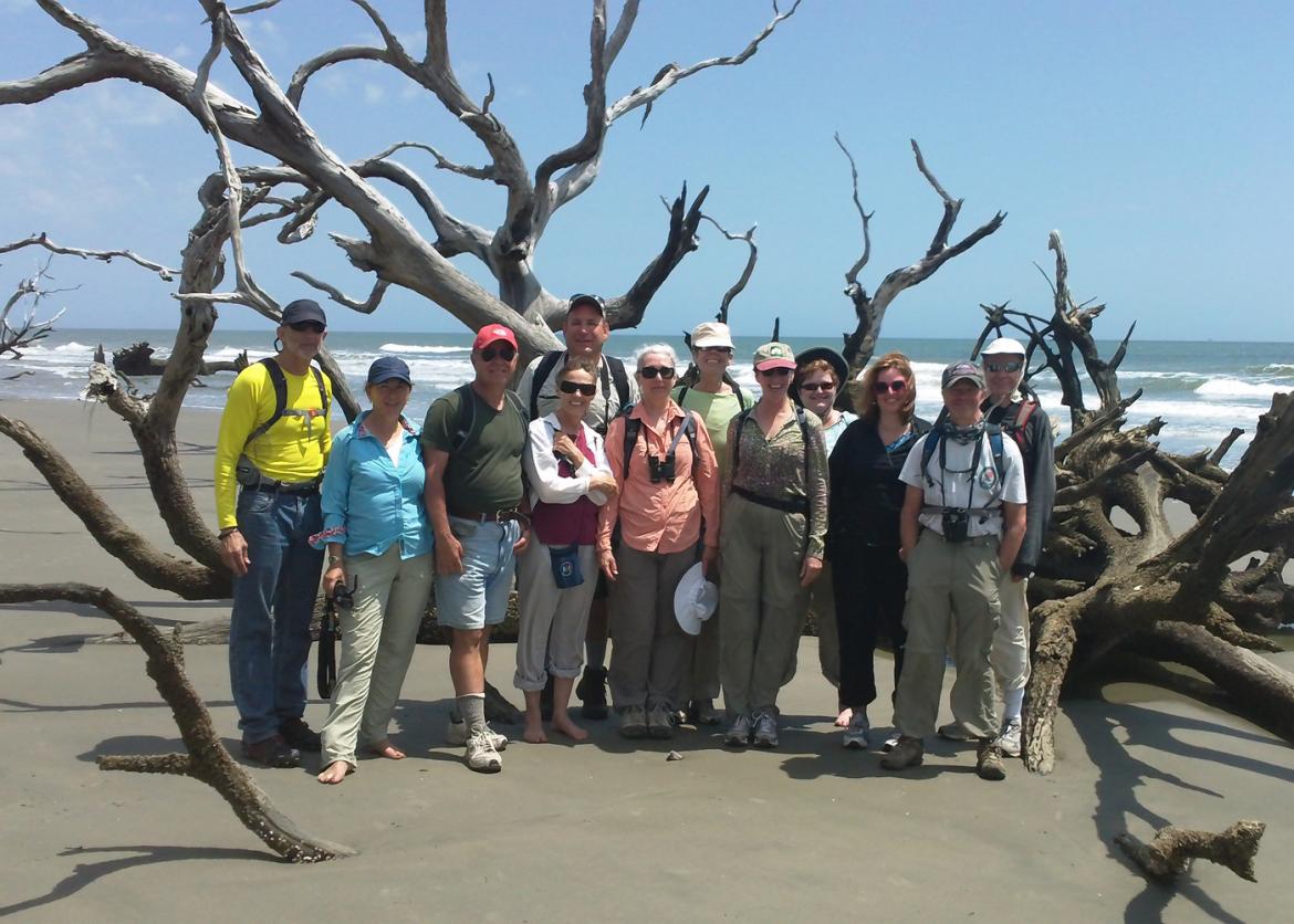 Twelve smiling people pose in front of a massive driftwood tree washed up on a beach.