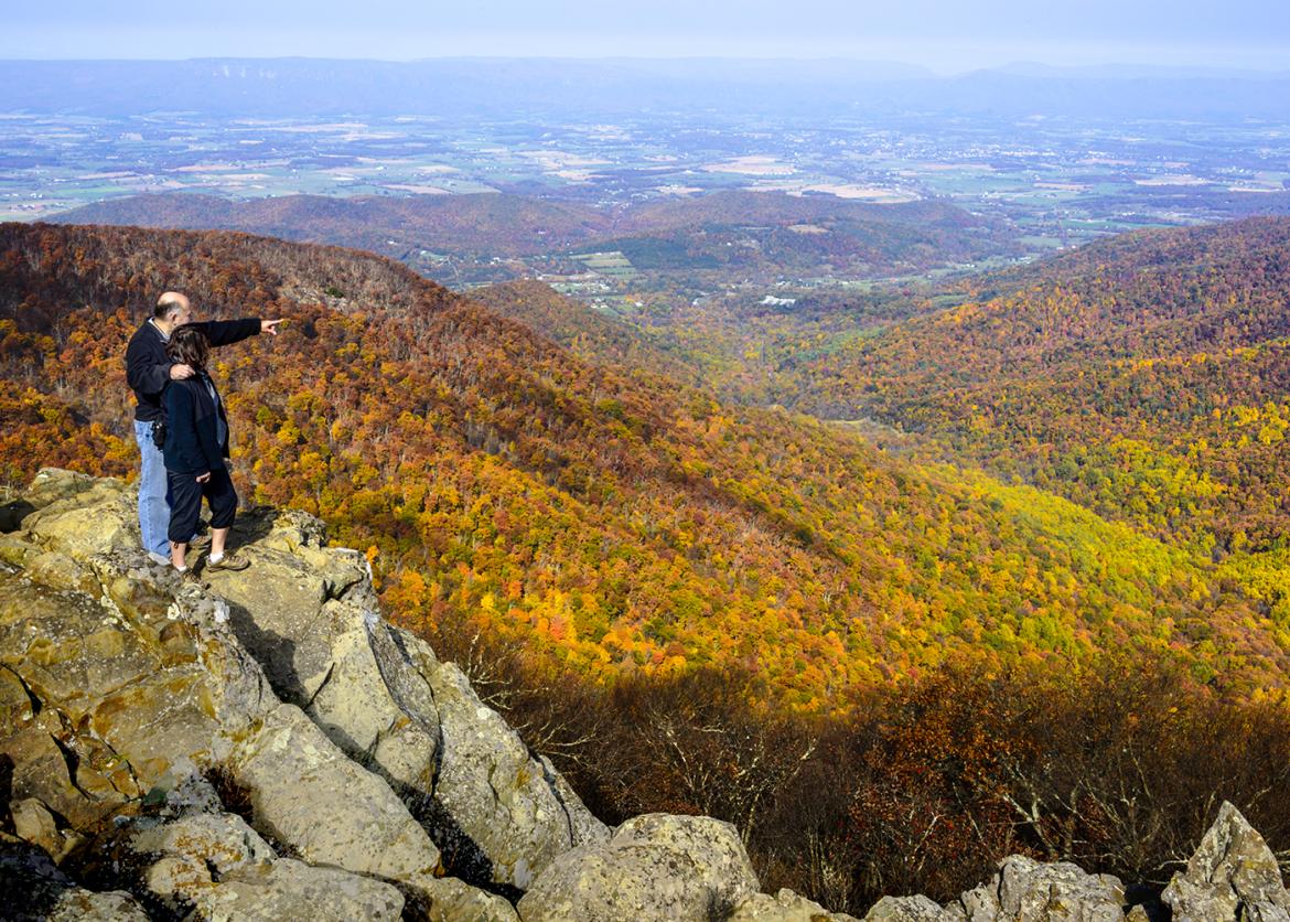 A couple stand on a scenic outlook. The man has his arm around the woman and points to the rolling slopes of autumn colored forest below.