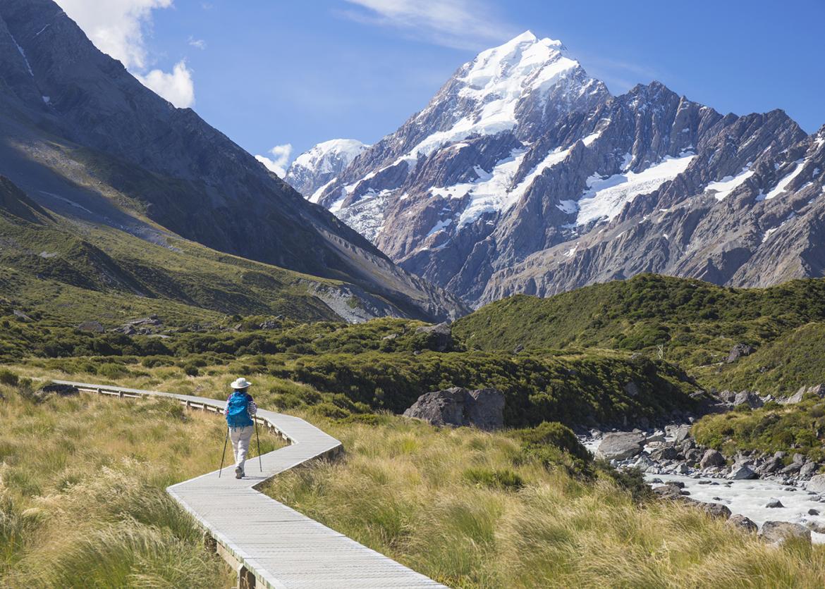A person hiking on a walkway over grass. A snowy mountain ridge stands in the distance.