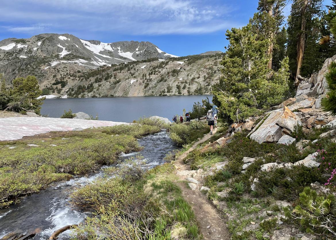 Hikers trekking towards a lake against a snow Sierra mountain backdrop