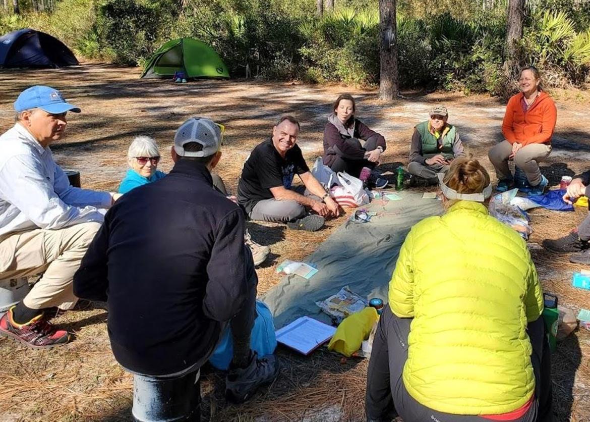Smiling people in outdoor gear sit and snack around a tarp at a camp site.