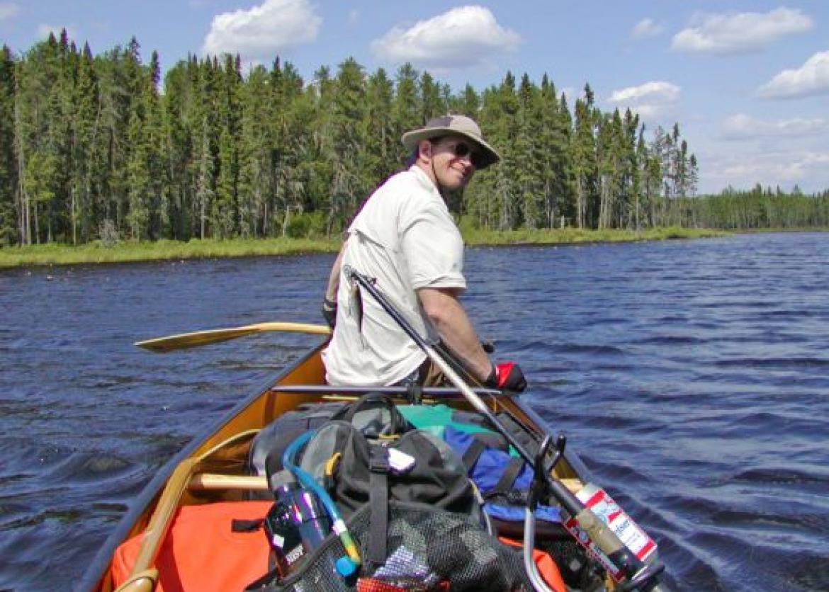 A man turned around, smiling on a canoe with the water and trees as the view.