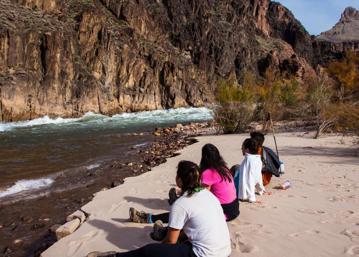 A group of people sitting on the sand next to the stream while looking at the canyon.