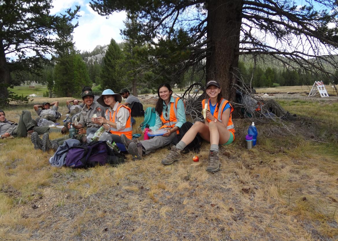 A group of smiling participants and park rangers sitting near trees eating lunch