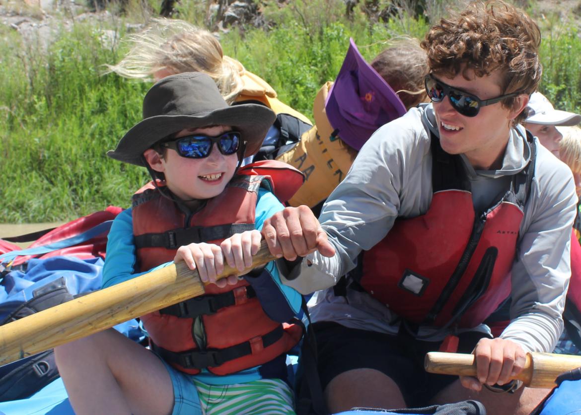 Two boys wearing sunglasses and life jackets holding oars on a crowded raft.  The older teenager is helping the younger kid hold his oar.