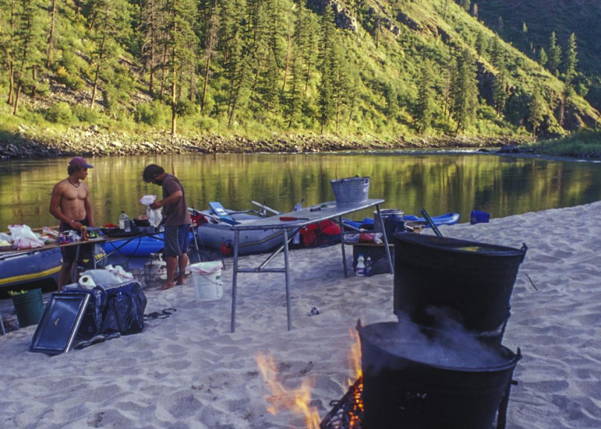 A camp set up on a sandy beach next to a lake and forested hills.  Two pots cook on a grill fire of coals and wood.