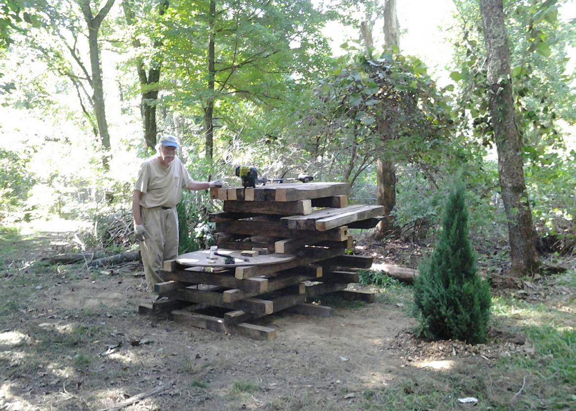 A man standing next to a pile of stacked wood with his hand on it.
