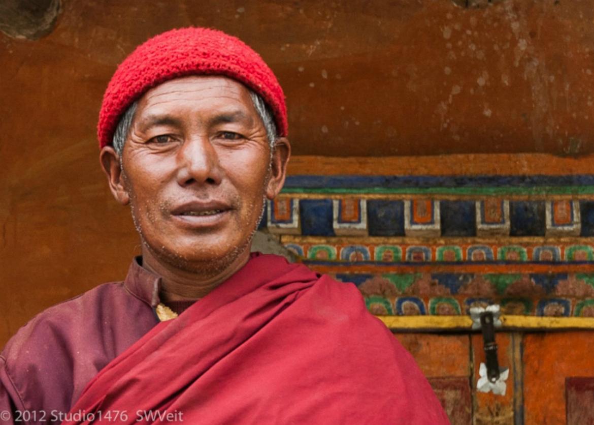A Nepalese man dressed in a red knit hat, tunic, and fabric wrap around the shoulder.  He stands in front of a painted door.