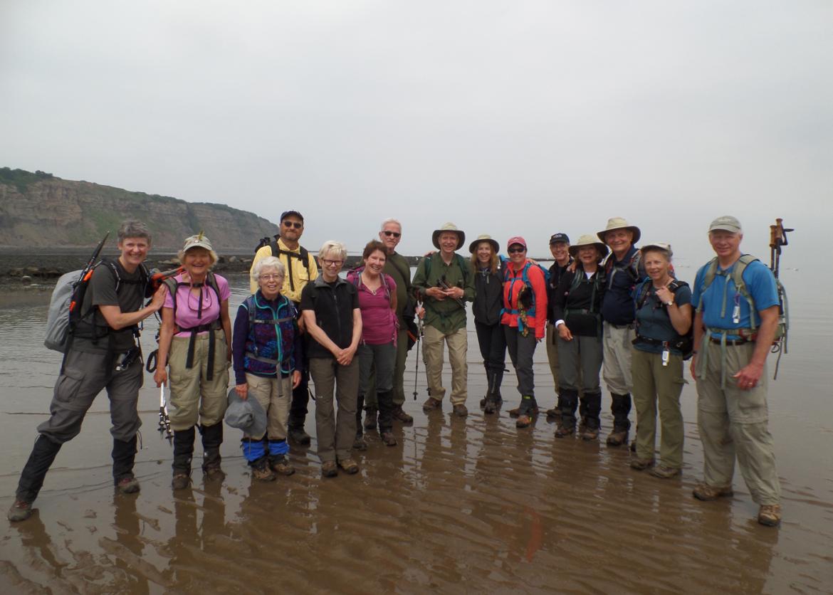 A group of fifteen smiling people in outdoor gear.  They are standing on wet brown sand.
