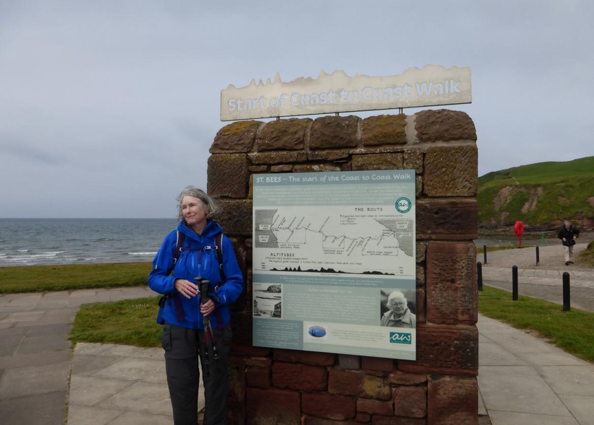 A woman stands next to a stone structure afixed with signs. The metal above reads "Start of the Coast to Coast Walk."  Another sign is informational sign and depicts a trail map.