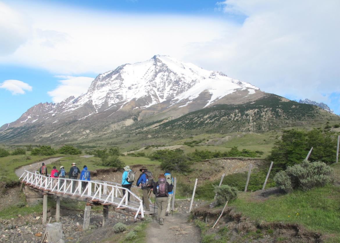 Eight people walk over a wooden bridge crossing a shallow gully. A snowy mountain stands in the distance.