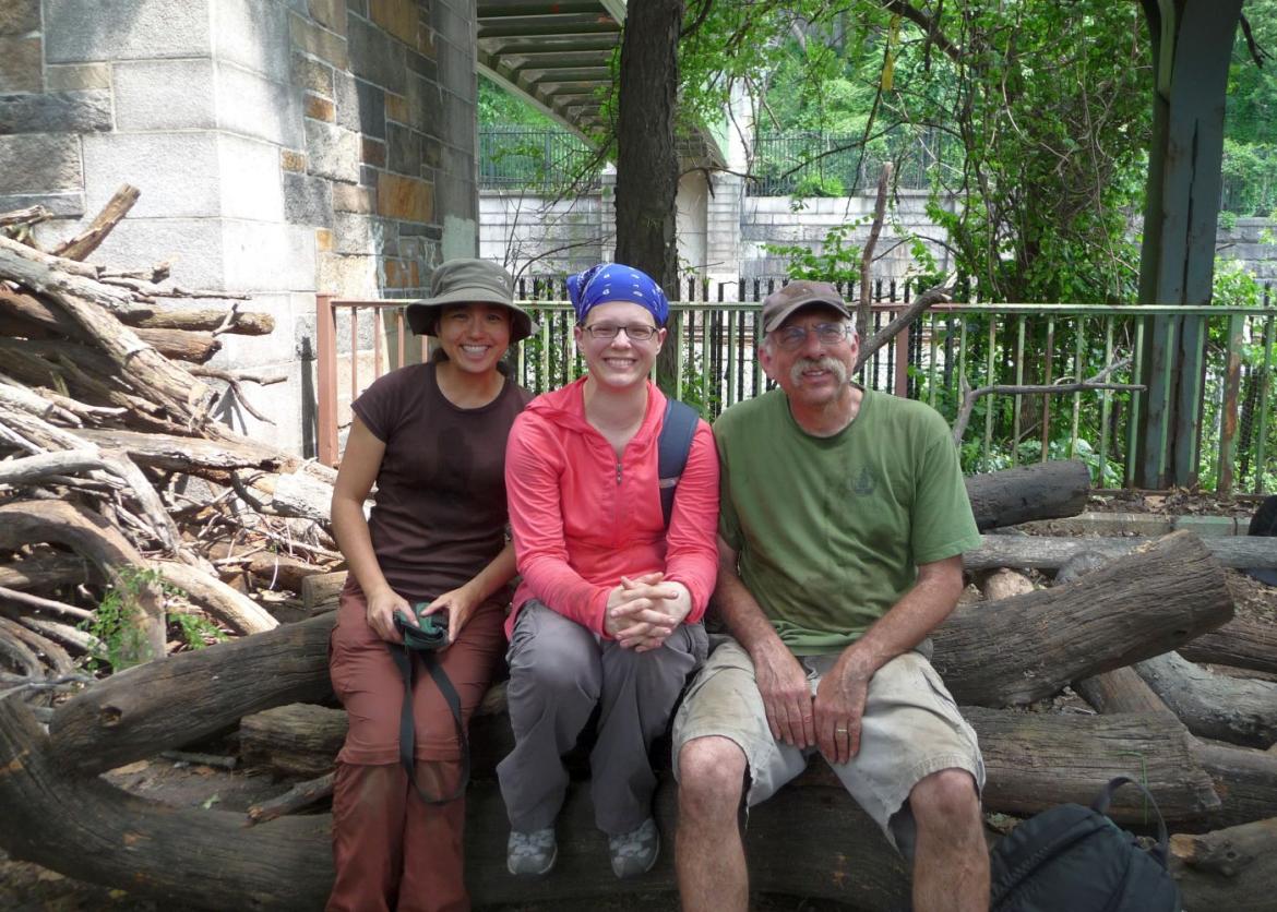 Three smiling people sitting on top of piled wooden tree logs.