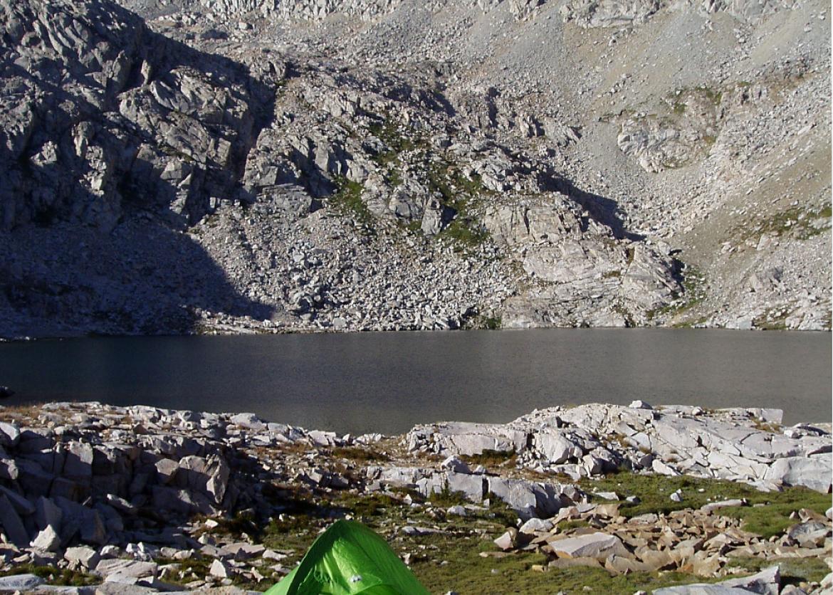 A green tent set up on the rocks next to the lake with the mountains in the background.