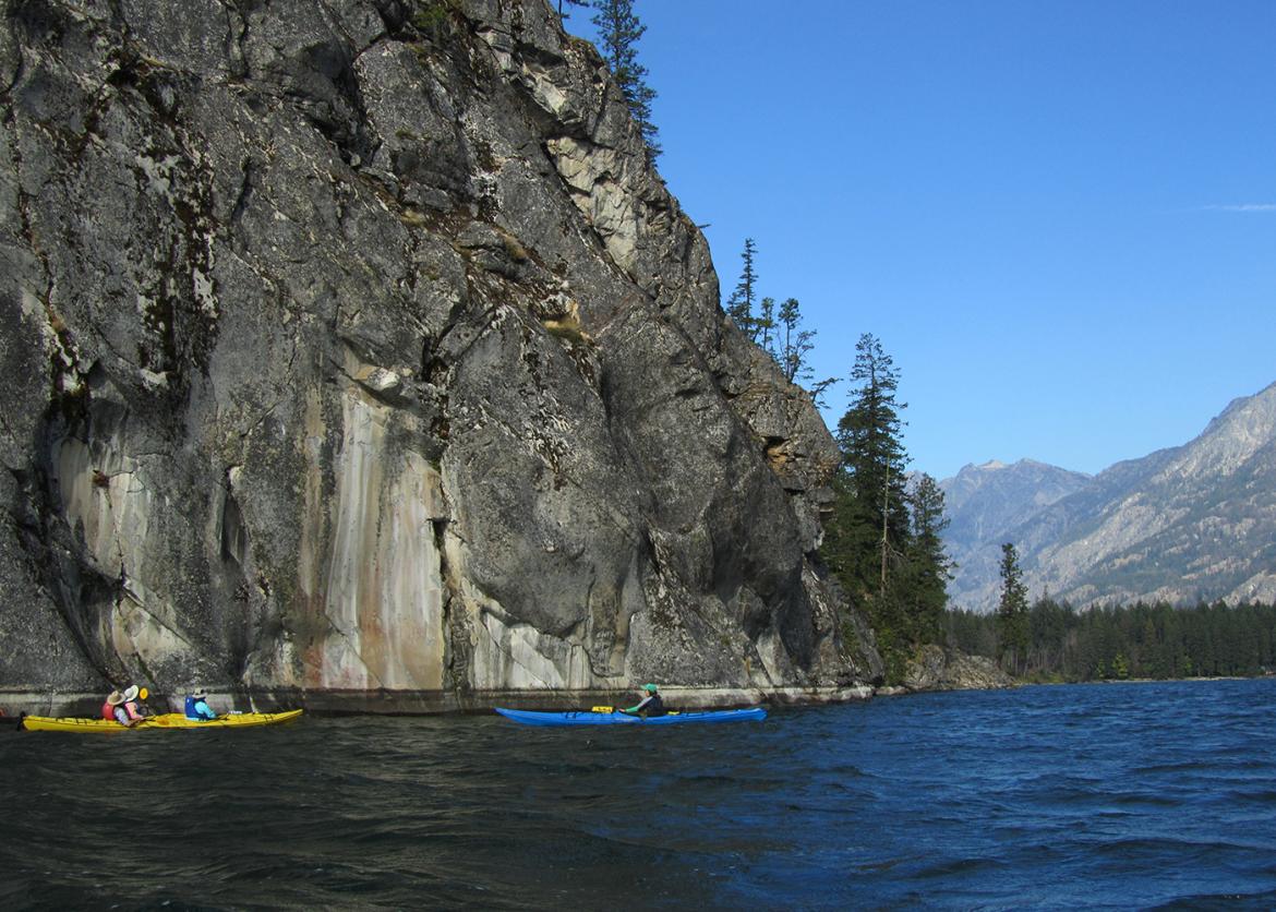 Kayakers paddle by a sheer rock face.