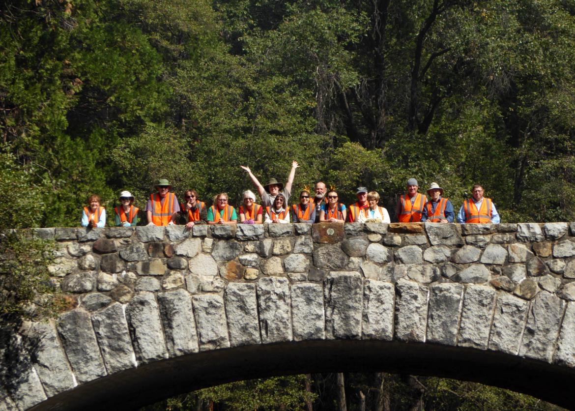 Smiling participants in work wear stand in a line on a stone bridge. One person in the center poses with their arms thrown in the air.