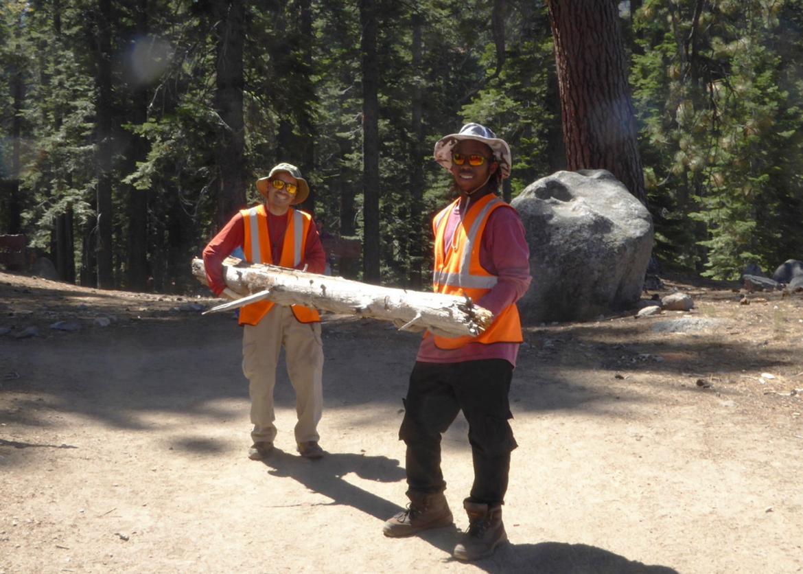Two smiling participants in work wear lift a tree log together.