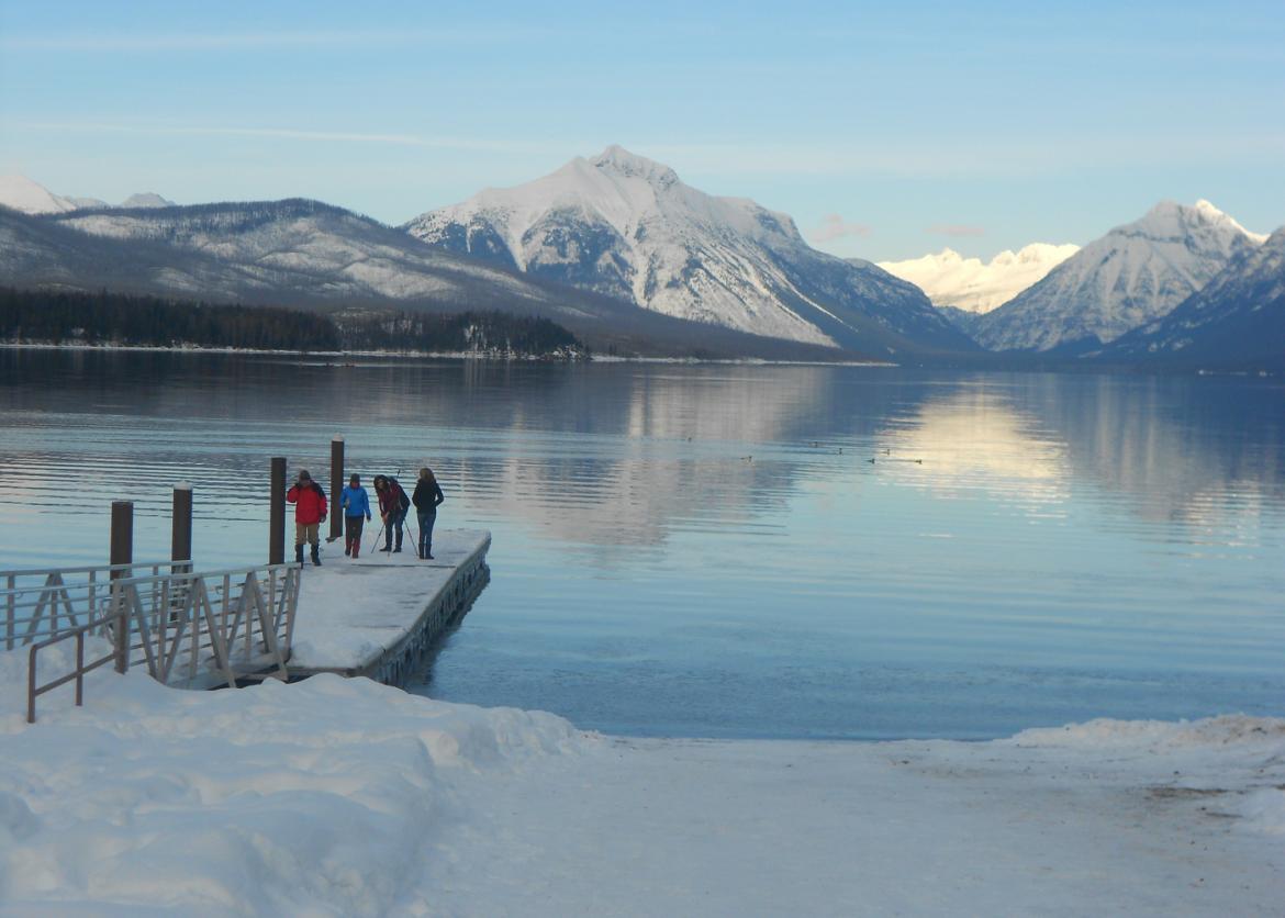 Four people on a snowy dock. On the otherside of the water are snowy mountains.