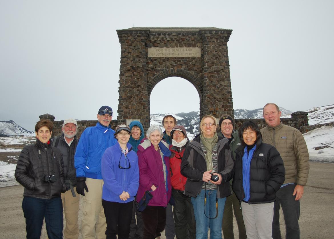 Twelve smiling people stand in front of a stone arch. The arch's engraving reads "For the benefit and enjoyment of the people."