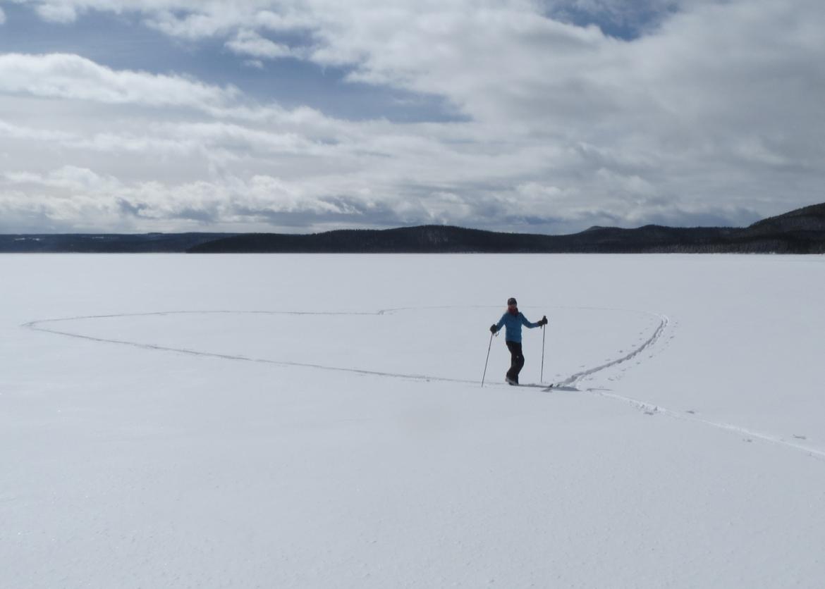 A skier poses for the camera, having finished carving a trail in the shape of a heart with their skis on flat ground.