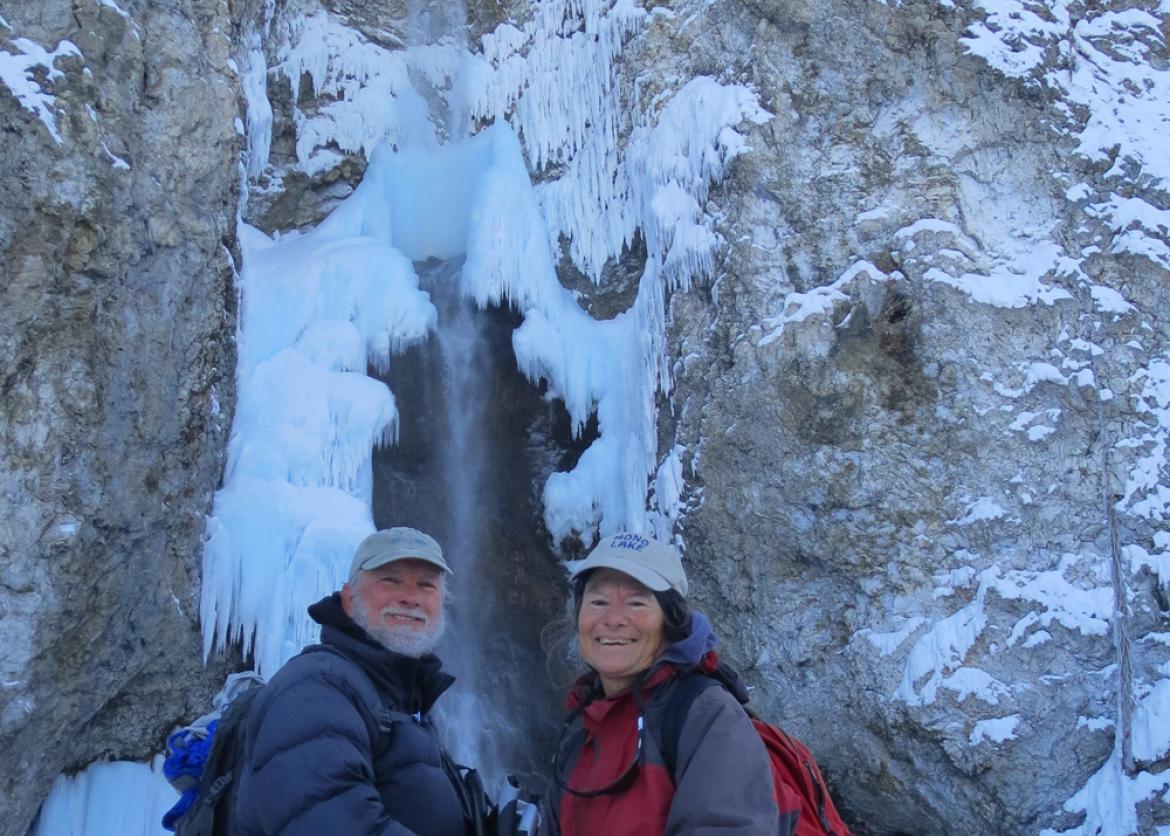 Two smiling people stand in front of the base of a frozen waterfall.