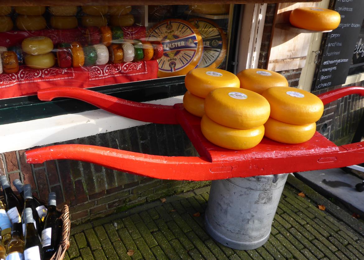Cheese wheels stacked on a red painted wooden sled.  The shop window display behind has even more cheeses.