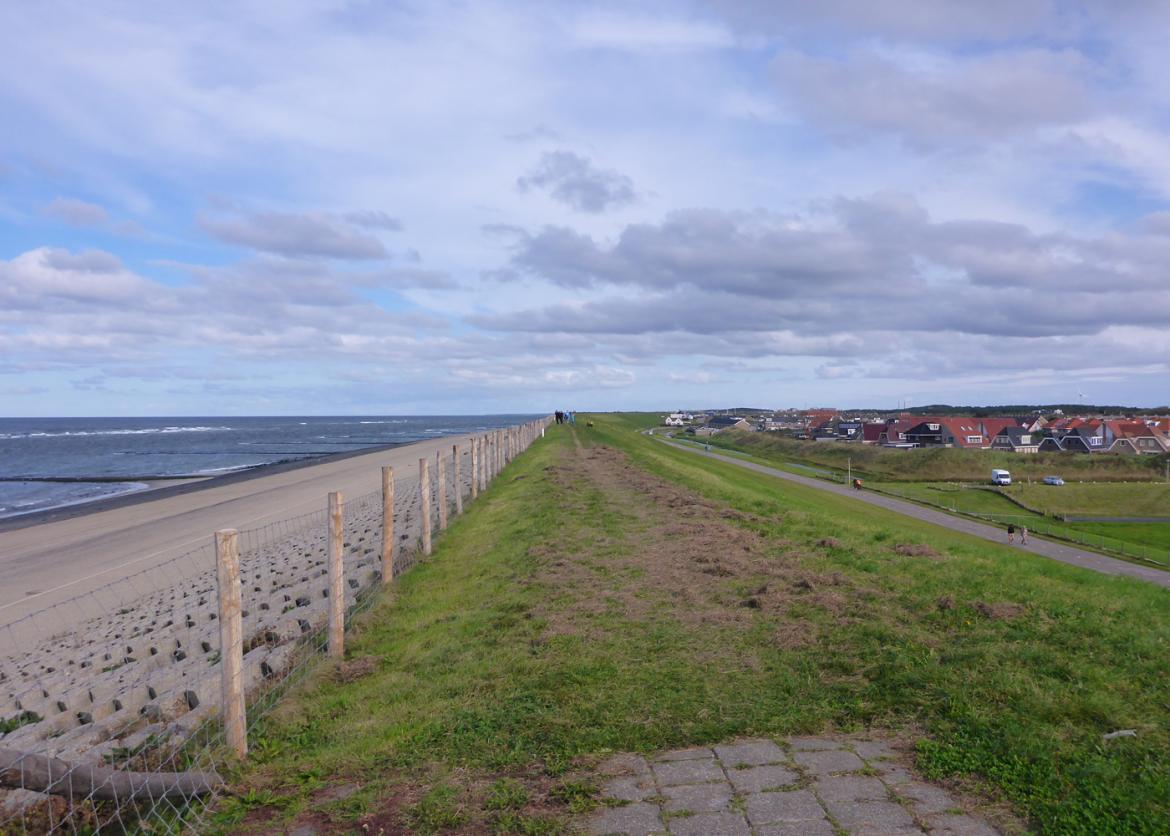 A long stretch of grass fenced off from the beach and water, with a town in the distance.