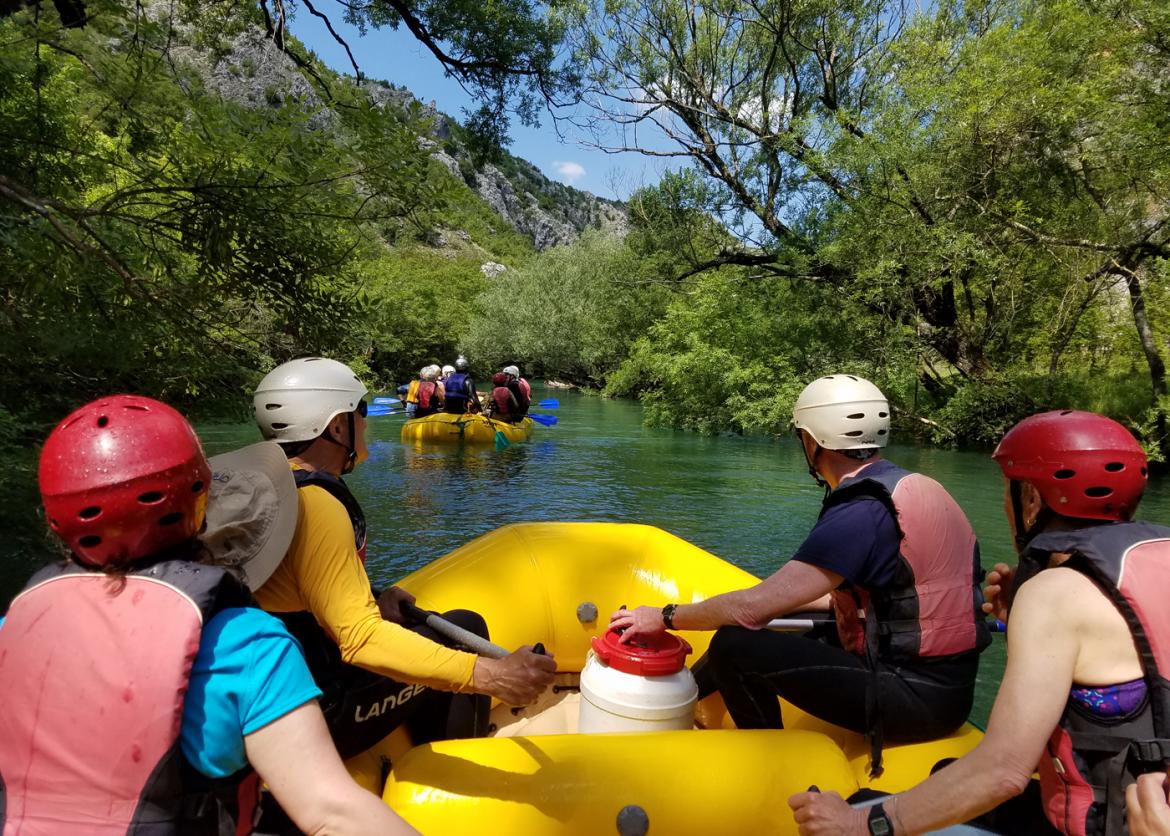 People sitting at the front of an inflatable raft and follow behind other rafts on a calm river.  The riverbanks are covered in trees.