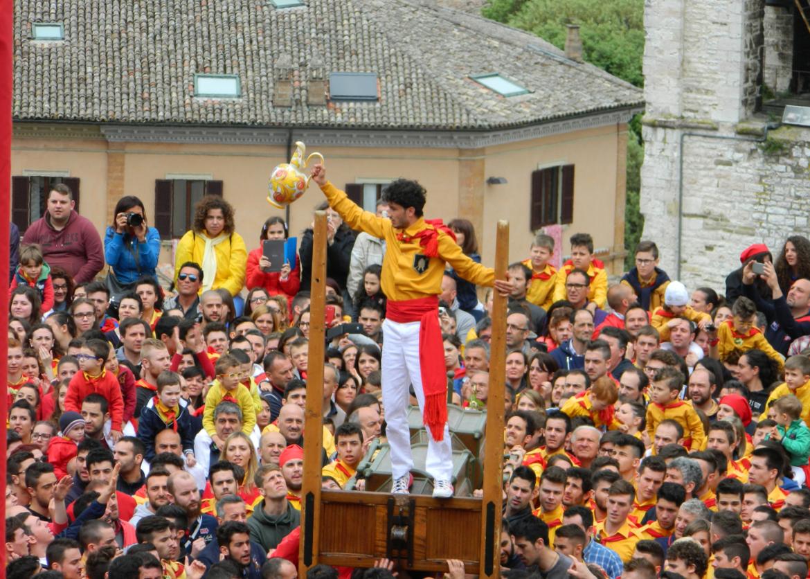 A man standing above a crowd on a wooden gate. He holds a decoorated ceramic pitcher aloft. He wears a red sasha around his waist.