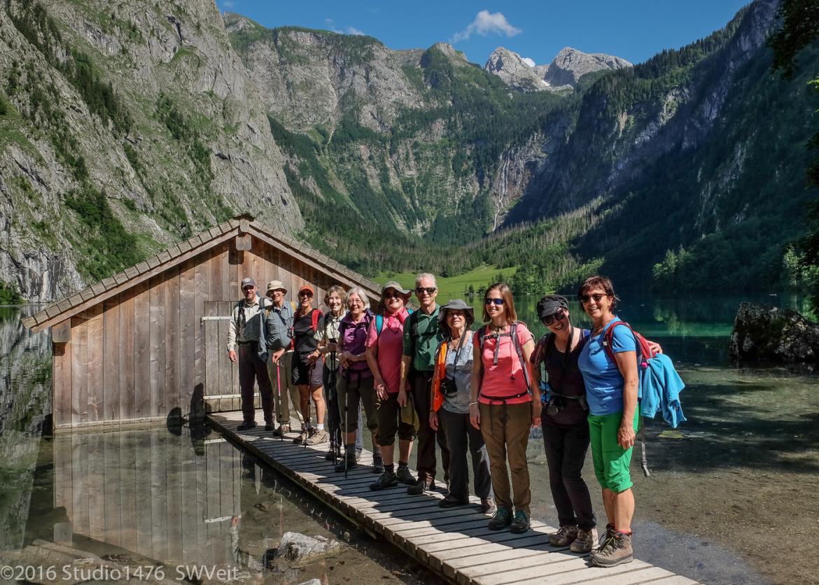Eleven smiling people stand single file on a wooden walkway leading up to a wood cabin door. The walkway crosses over a pooled puddle of water.