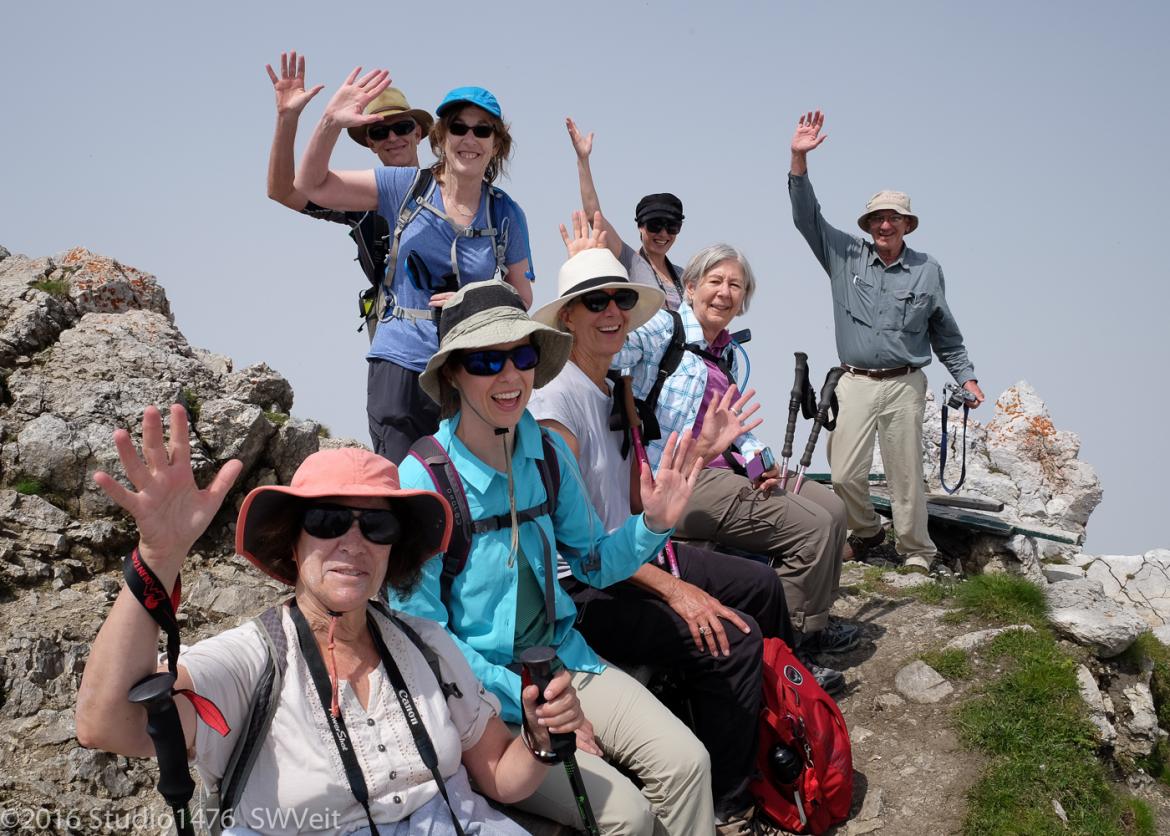 Hikers smile and wave at the camera from where they're seated on a rock.