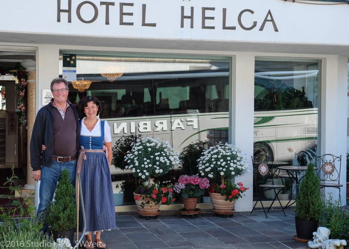 A couple with their arms around each other stand in front of a building which is titled with the words "Hotel Helga." A bus is reflected in the windows.