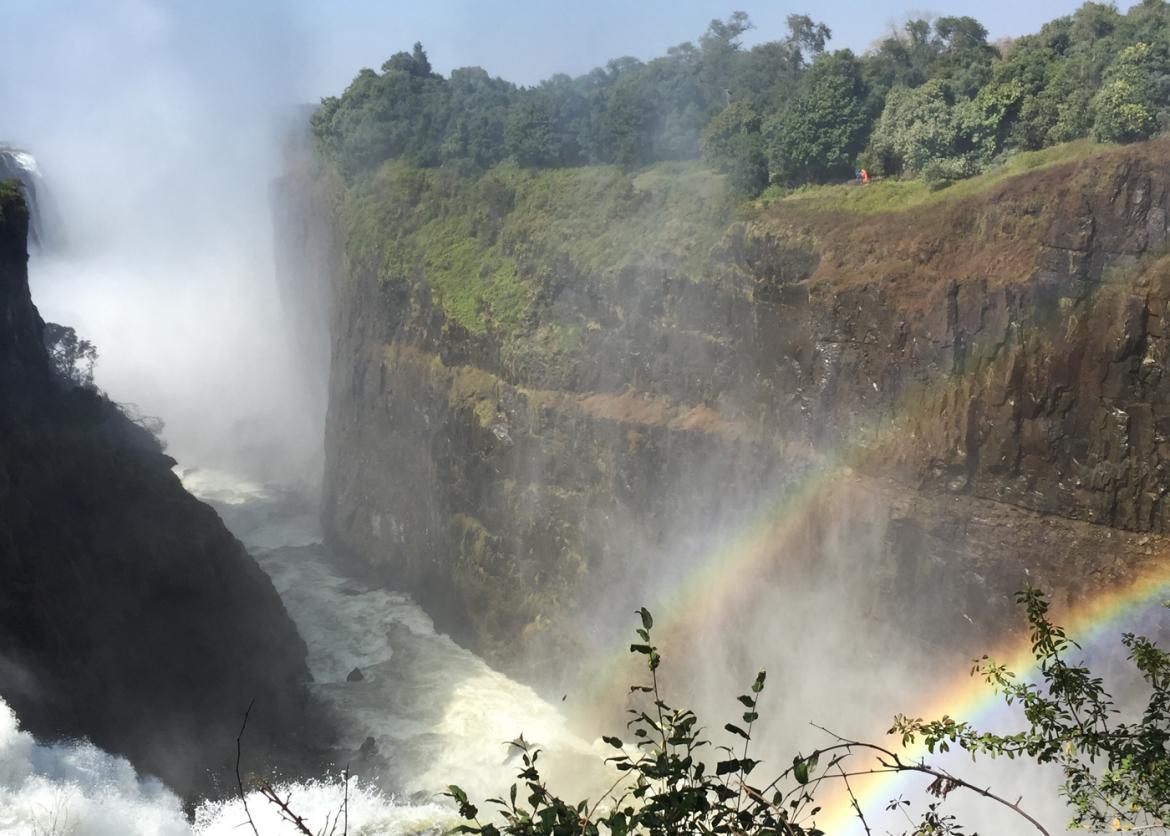 Rainbows over a misty river flowing through a canyon.  One of the sheer canyon walls is topped by trees.