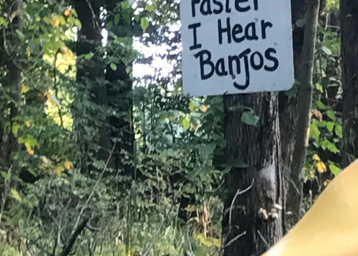 A view over a kayak paddle of a sign nailed to a tree that reads "Paddle faster I hear banjos."