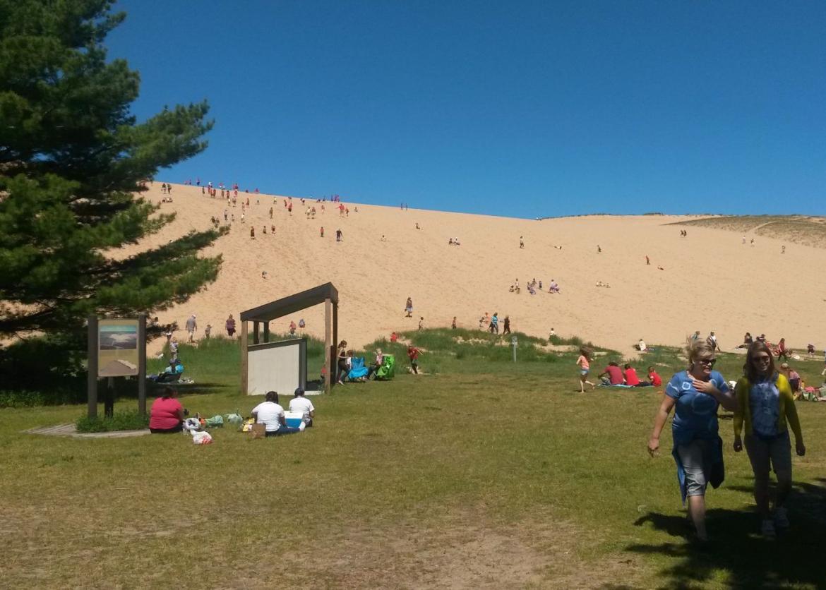A park with a large, green field and a sandy area above the hill filled with people relaxing and chilling.