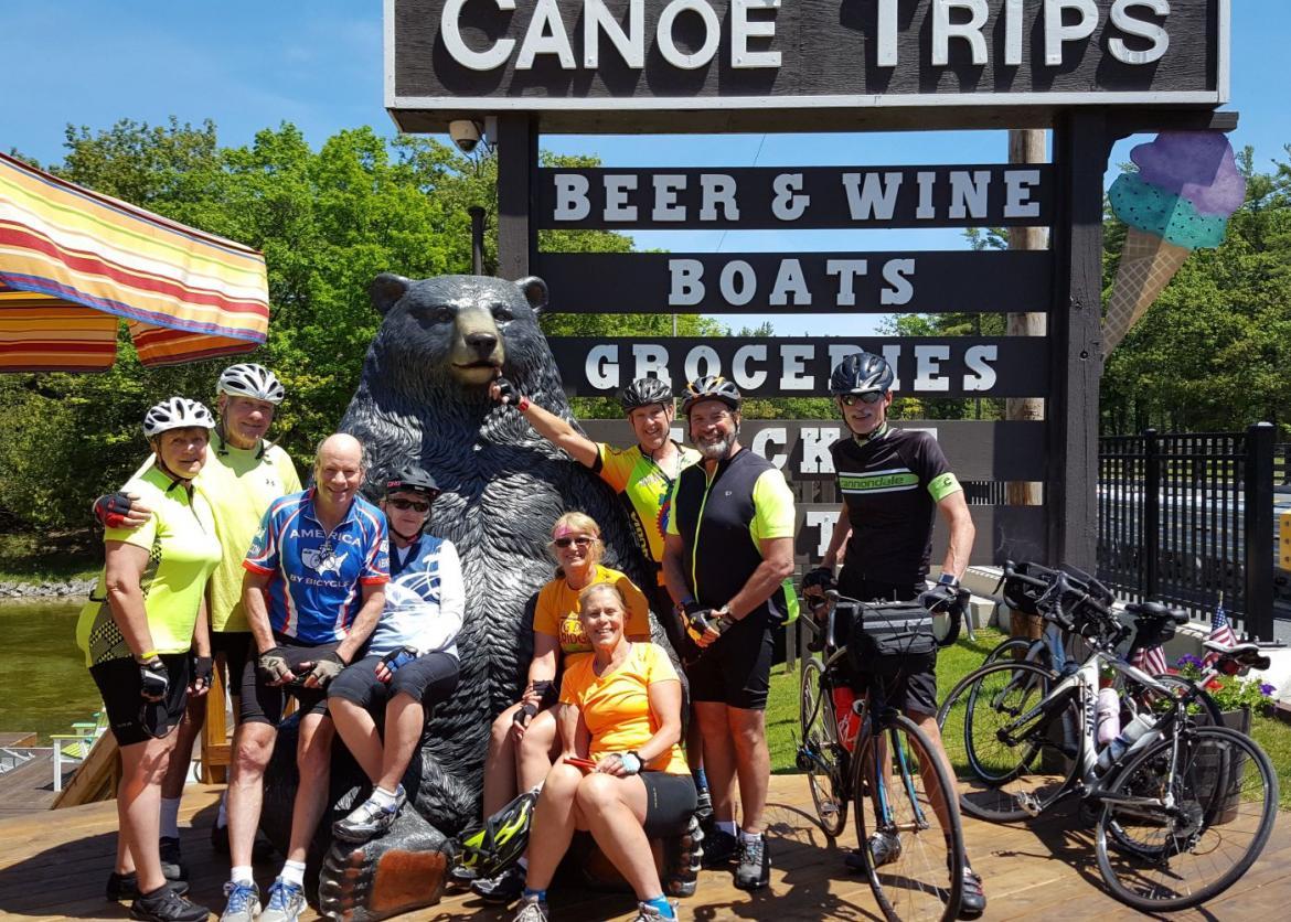 A group posing and pointing at the bear statue and sign that says "Beer & Wine Boats Groceries".