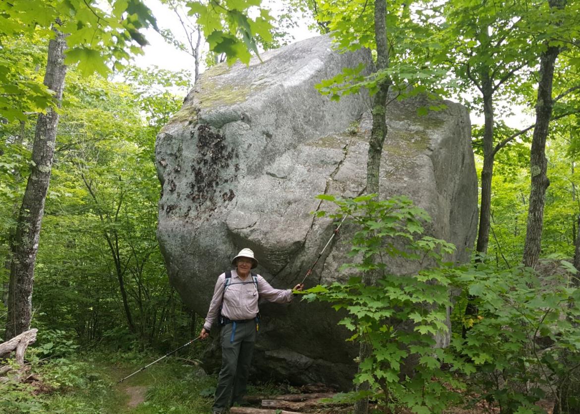 A man posing as he holds up his trekking pole in front of a large rock.