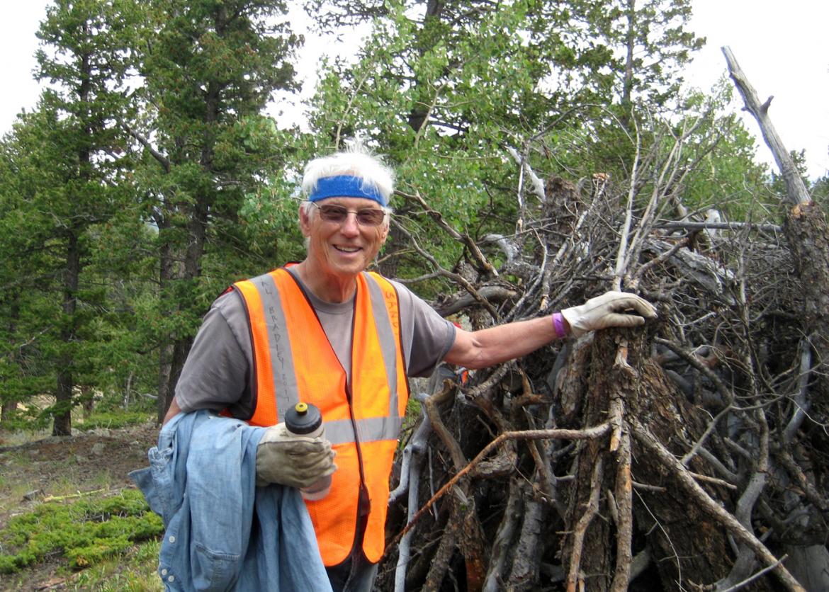A man in a high visibility vest rests his arm on a pile of gnarled dead tree branches. The pile stands as tall as he is.
