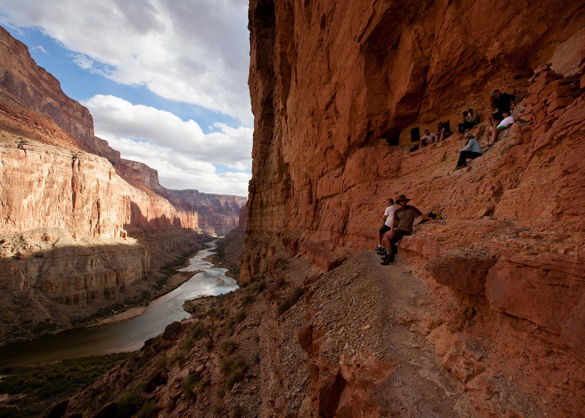 Several people sit in various place on an indent in a canyon wall.  The shadow of the canyon wall covers the people and stretches to the opposite wall.