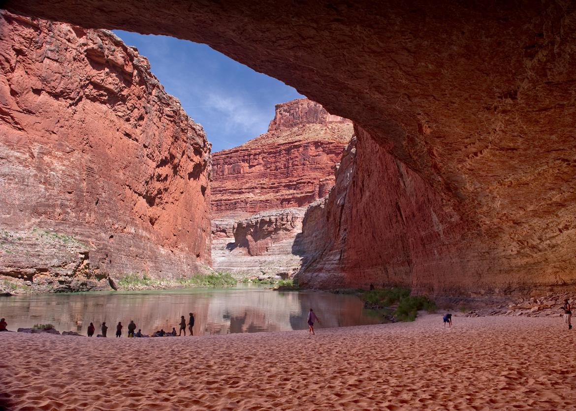 A massive naturally formed canopy of rock arches over several people who gather in the sand at the water's edge.
