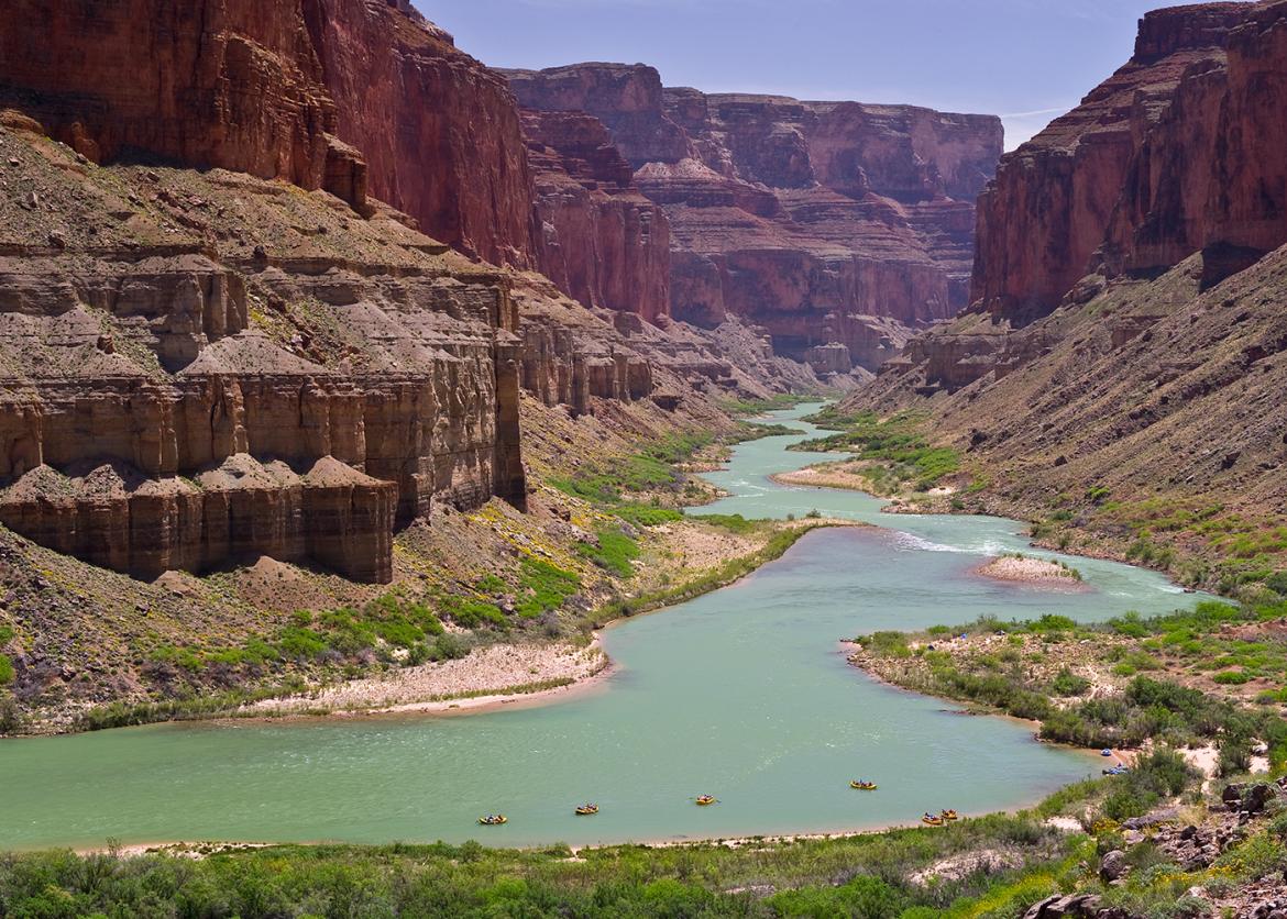 A river flows through a massive canyon.  In the distance, several rafts head downstream.