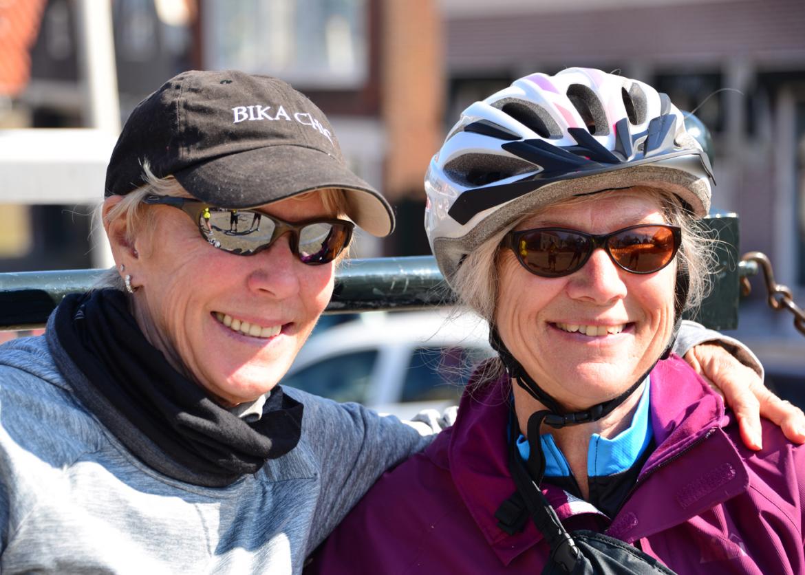 Two smiling women in sunglasses.  One in a baseball cap has her arm around the other. The other woman is wearing a bike helmet.