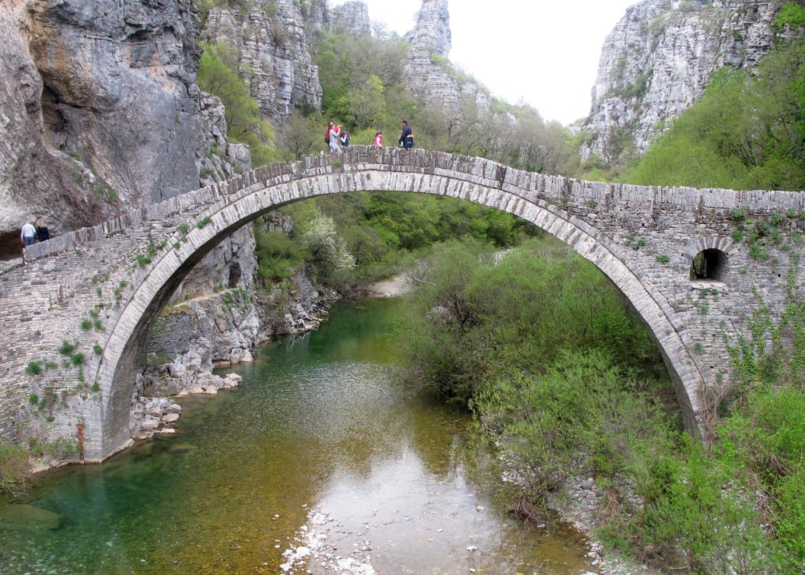 People walking over river on a stone arch bridge inside a canyon.