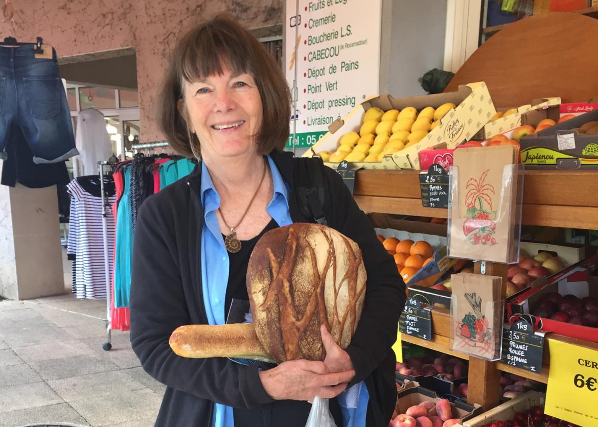 A woman holding two loaves of bread in front of a grocery stand full of fruit.