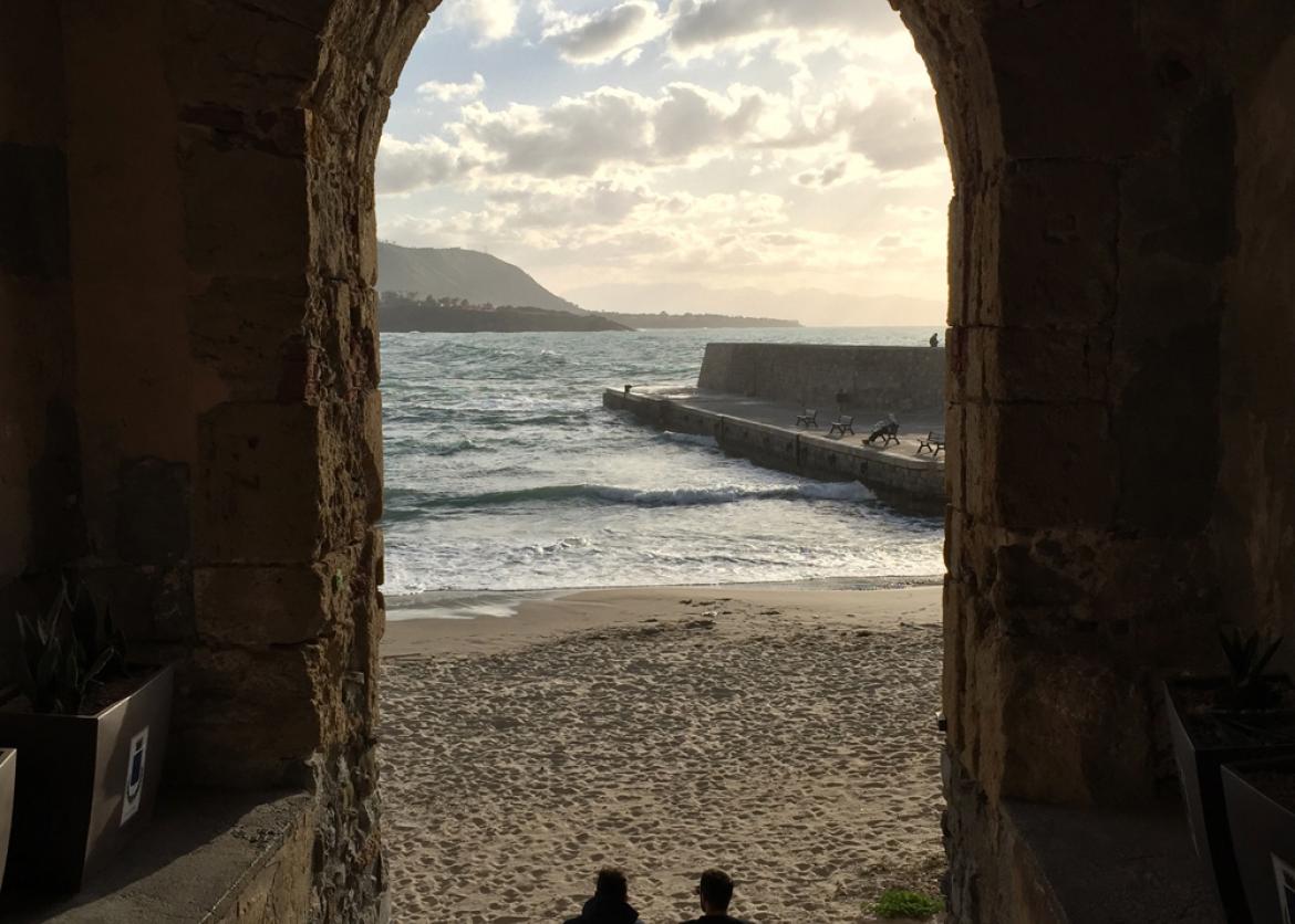 Two people sit under a tall stone arch, overlooking a beach.
