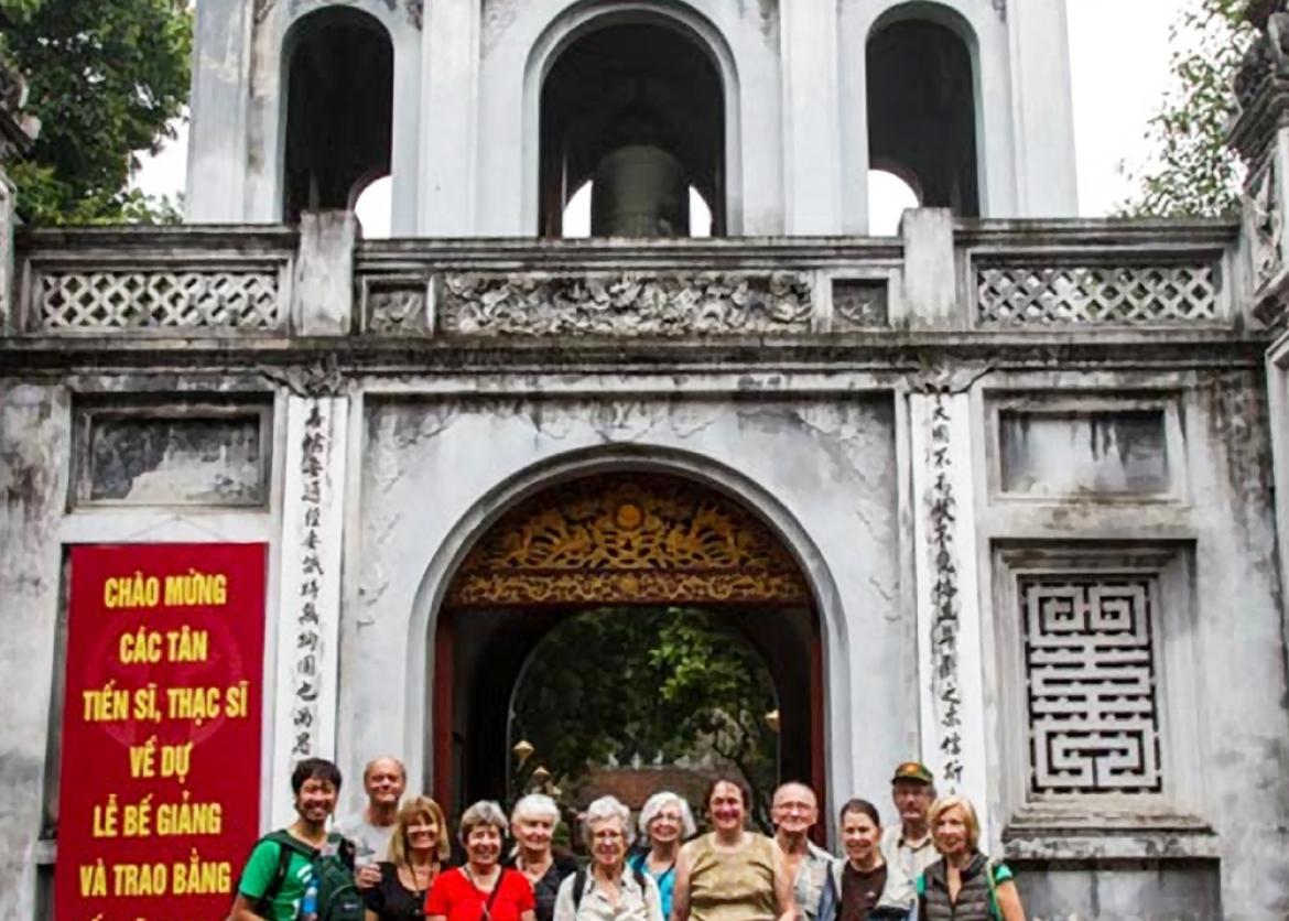 Sixteen smiling people pose in front of an ancient stone building.