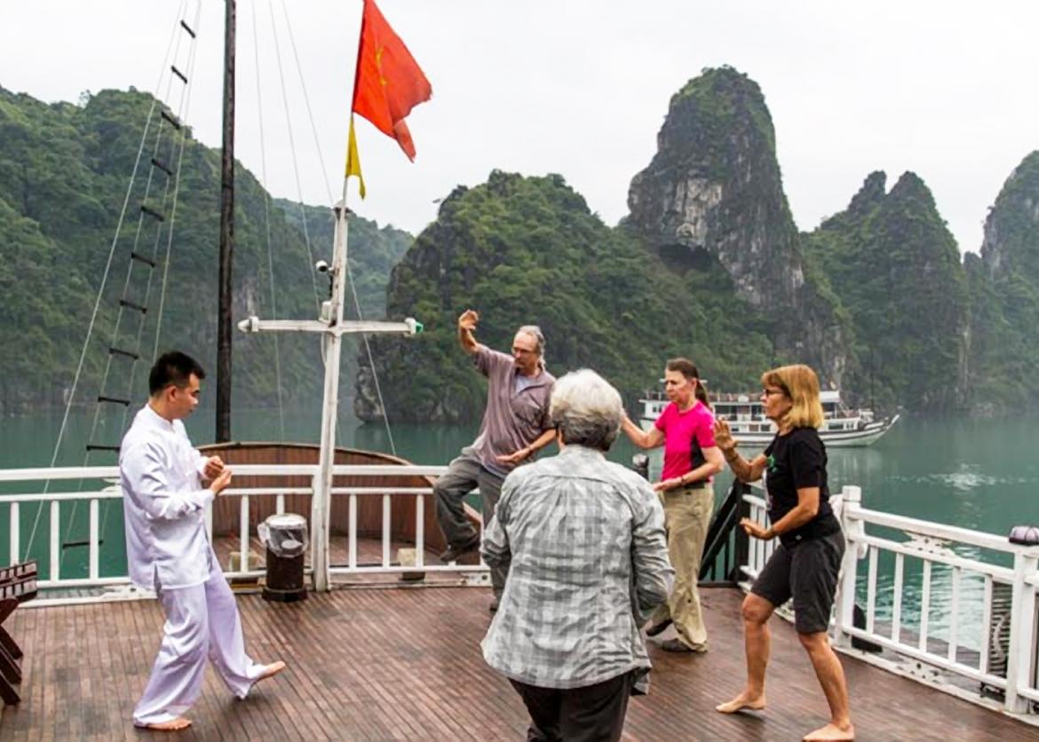 On the deck of a boat, three people sweep their arms, the movement mimicking their traditionally dressed Vietnamese instructor.