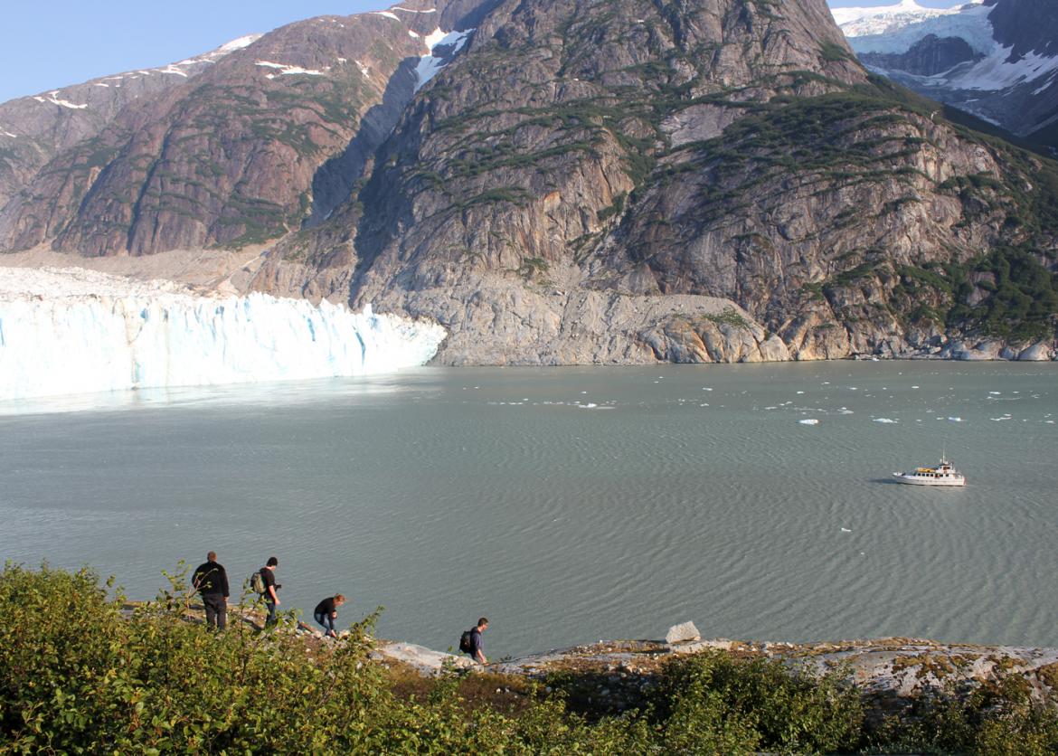 A view of a body of water, a glacier cliff, a snow and greenery covered mountain.  Four people look on from a cliff edge, and a boat floats in the water.