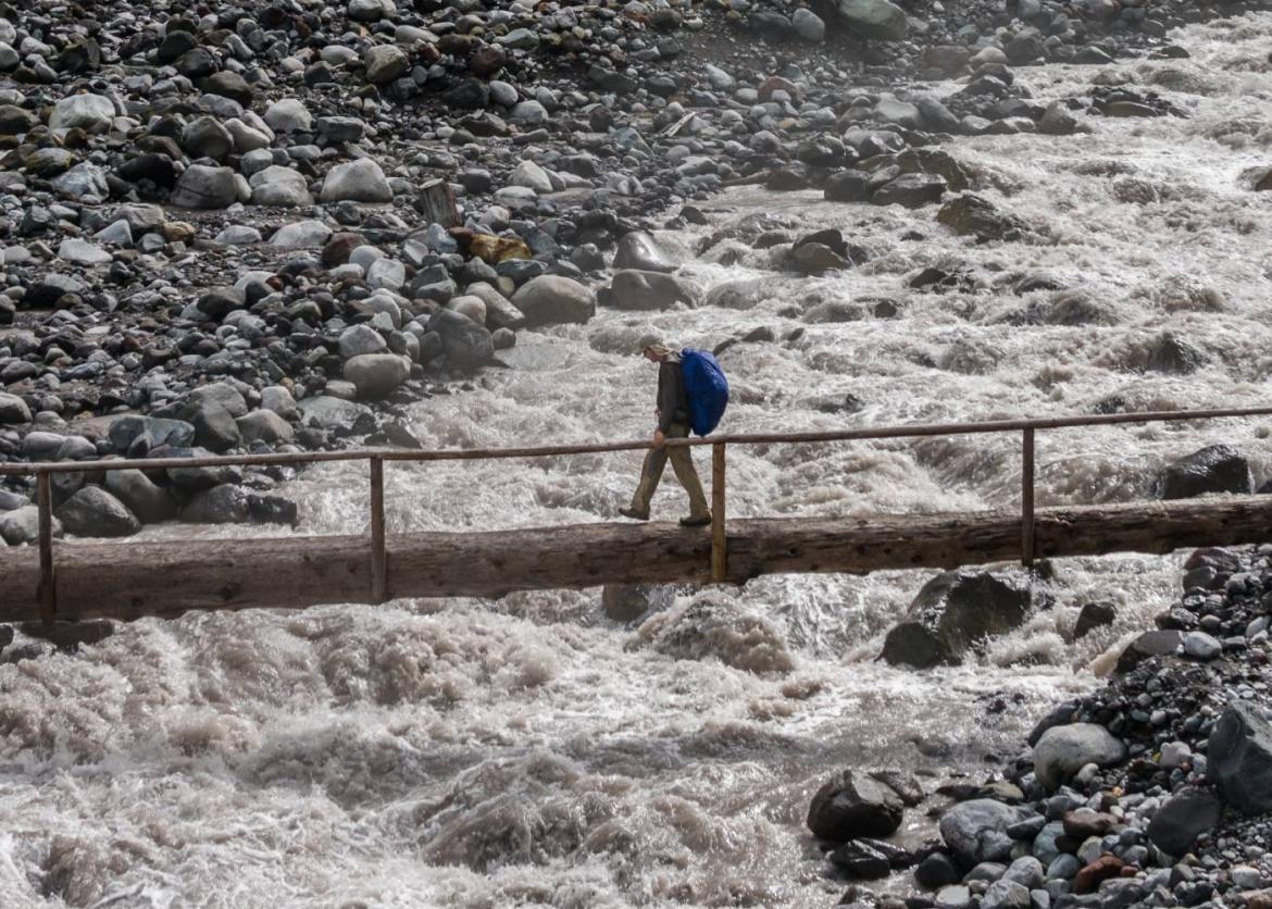 A person walks over a bridge. The bridge is a single tree trunk laid on its side with a wooden railing attached on one side. White water rushes beow.