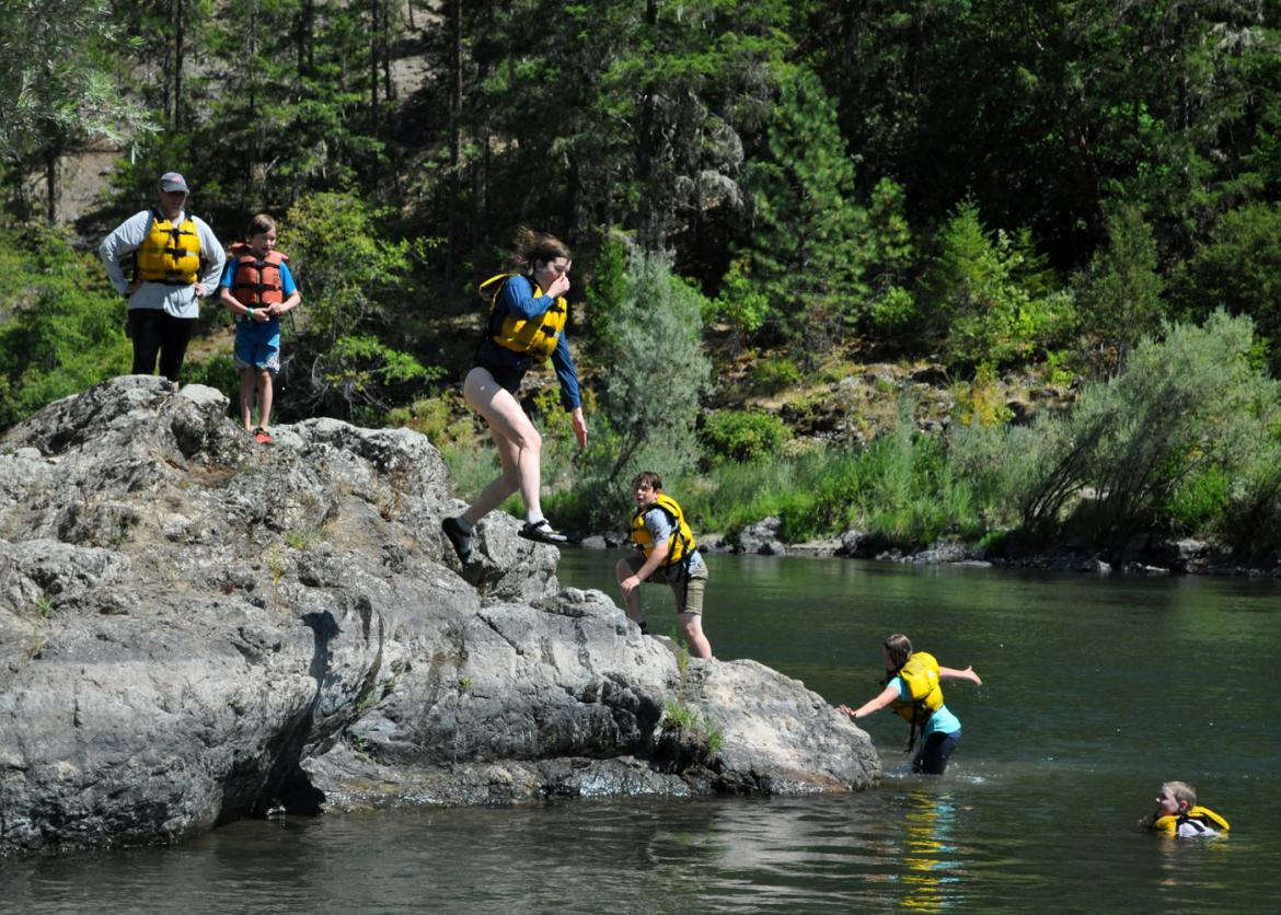 A girl in a life jacket holds her nose as she jumps into the water. Other youths in life jackets climb out of the water and onto a rock.